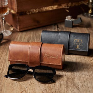 May include: A brown leather eyeglasses case with the name "Michael" in gold script, next to a pair of black sunglasses. A black leather case with the name "TASTY" is also visible. The cases are on a wooden surface.