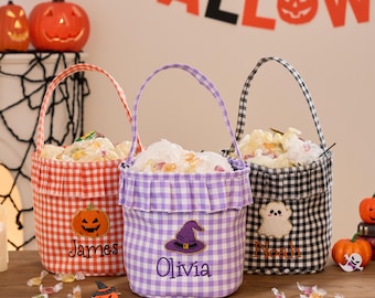 May include: Three fabric Halloween candy buckets with gingham patterns in red, purple, and black. Each bucket features a ruffled top, a handle, and an embroidered design: a pumpkin for James, a witch hat for Olivia, and a ghost for Noah.