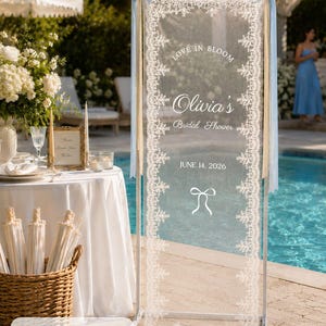 K&ouml;nnte beinhalten: Ein vertikales rechteckiges Schild mit einer transparenten wei&szlig;en Stoffbahn mit aufwendigen Spitzenr&auml;ndern, aufgeh&auml;ngt in einem wei&szlig;en Metallrahmen. Der Text lautet LOVE IN BLOOM, Olivia's Bridal Shower, JUNE 14, 2026, mit einem Schleifensymbol. Ma&szlig;e 75 x 200 cm.