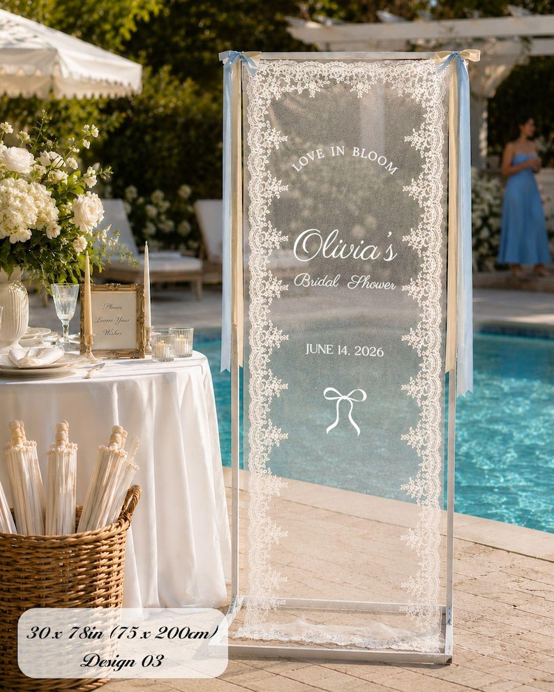 K&ouml;nnte beinhalten: Ein vertikales rechteckiges Schild mit einer transparenten wei&szlig;en Stoffbahn mit aufwendigen Spitzenr&auml;ndern, aufgeh&auml;ngt in einem wei&szlig;en Metallrahmen. Der Text lautet LOVE IN BLOOM, Olivia's Bridal Shower, JUNE 14, 2026, mit einem Schleifensymbol. Ma&szlig;e 75 x 200 cm.