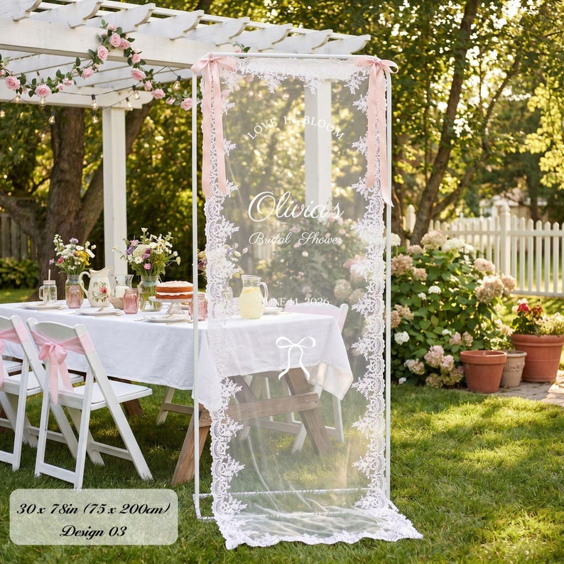 K&ouml;nnte beinhalten: Ein wei&szlig;er Metallrahmen mit einem transparenten, spitzenbesetzten Stoffpaneel, bedruckt mit dem Text "LOVE IS BLOOM", "Olivia's", "Bridal Shower" und "JUNE 2026". Das Paneel zeigt eine Schleifengrafik, misst 75 mal 200 cm und ist mit rosa B&auml;ndern verziert.