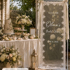 K&ouml;nnte beinhalten: Ein hoher rechteckiger wei&szlig;er Metallrahmen mit einem transparenten Spitzenpaneel, das den Text Welcome to Olivia's Bridal Shower June 14, 2026 tr&auml;gt. Die Spitze hat florale R&auml;nder und misst laut Produktetikett 75 x 200 cm.