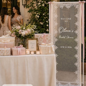 K&ouml;nnte beinhalten: Ein hohes, rechteckiges Hochzeitsschild mit den Ma&szlig;en 75 x 200 cm, das einen wei&szlig;en Spitzenrand auf einer transparenten Platte aufweist. Der Text lautet: WELCOME TO Olivia's Bridal Shower JUNE 14, 2026. Es ist wei&szlig; gerahmt und mit rosa B&auml;ndern verziert.