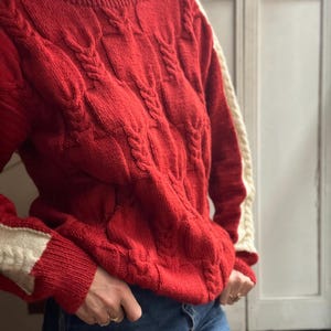 Könnte beinhalten: Ein roter Zopfstrickpullover mit cremefarbenen Akzenten an Schultern und Ärmeln. Der Pullover hat einen Rundhalsausschnitt und ein strukturiertes, gewebtes Design. Die Ärmel und Schultern haben eine kontrastierende cremefarbene Farbe.