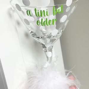 May include: A clear glass martini glass with white polka dots and the text "a tini bit older" in green lettering. The glass is sitting on a white fluffy pom-pom.