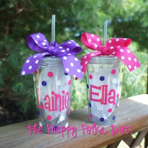 May include: Two clear plastic cups with pink polka dots and blue polka dots. Each cup has a straw and a purple or pink bow. The cups are personalized with the names "Ella" and "Laine".