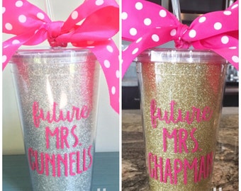 May include: Two clear plastic cups with gold and silver glitter. The cups have pink polka dot bows and straws. The text "Future Mrs." is written in pink on each cup, followed by a last name.