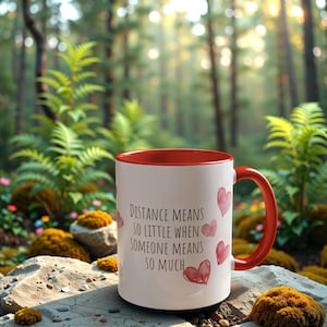 Puede incluir: Taza de cerámica blanca con borde y asa rojos. La taza tiene una cita que dice "La distancia significa tan poco cuando alguien significa tanto." con corazones rosas de acuarela.