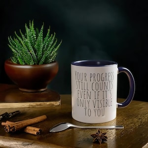 Puede incluir: Taza de café blanca con borde y asa morados. La taza tiene el texto "Your progress still counts even if it's only visible to you" impreso en negro.