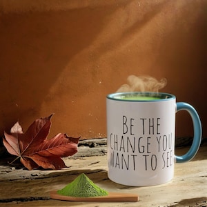 Puede incluir: Taza de cerámica blanca con borde y asa azules. La taza tiene el texto "Be the change you want to see" impreso en ella. La taza está llena de una bebida verde y sale vapor de ella. En primer plano, hay un pequeño montón de polvo verde en una cuchara de madera.