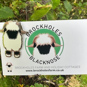 May include: A Blacknose sheep enamel pin with a cream body, black face, and brown horns, displayed with a card. The card features a sheep graphic and the text "BROCKHOLES BLACKNOSE FARM AND HOLIDAY COTTAGES".
