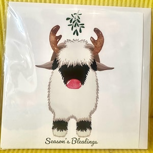Könnte beinhalten: Eine Grußkarte mit einer Cartoon-Yak-Illustration. Das Yak hat braune Hörner, ein schwarzes Gesicht und eine rote Nase. Es hat weißes Fell und schwarze Hufe. Der Text "Season's Bleatings" ist unten aufgedruckt, mit Mistel über dem Yak.