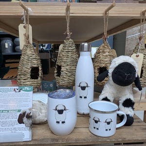May include: A selection of sheep-themed items, including a white water bottle, a tumbler, and a mug, each with a sheep illustration. A stuffed sheep toy and woven birdhouses are also present. A sign provides information about the sheep.
