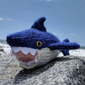 Peut inclure: Un jouet en peluche de requin bleu tricoté avec des dents blanches et un œil jaune, assis sur un rocher gris avec l'océan en arrière-plan.