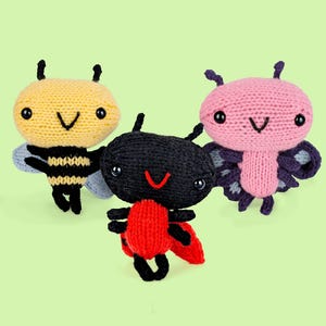 May include: Three small, handmade knitted plush toys shaped like insects. One is a yellow and black bee, one is a black and red ladybug, and one is a pink and purple butterfly. Each toy features black bead eyes and embroidered mouth details.