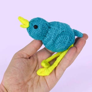 May include: A handmade knitted bird toy in shades of teal and yellow. The bird has a round body, a yellow beak, and yellow legs. The toy is held in a hand against a light purple background. The bird's eyes are black.