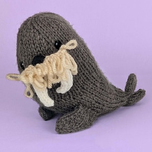 May include: A dark gray knitted walrus plush toy featuring a textured cream-colored yarn mustache and two prominent white tusks. The stuffed animal has small black bead eyes and a black nose, with visible knit stitches throughout its body.