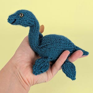 May include: A small, hand-knitted plush toy shaped like a long-necked sea creature, crafted from dark teal yarn. The creature features a rounded body, four flipper-like appendages, and a long neck with a small head containing a single visible dark eye.