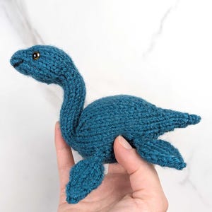 Op de afbeelding: Een tealblauw gebreid pluche speelgoed in de vorm van een plesiosaurus, een langhalzige zeereptiel. Het speelgoed heeft een klein, rond hoofd met zwarte ogen en een lange, slanke nek.