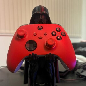 May include: A red Xbox controller is held by a Darth Vader figurine. The figurine is standing on a black base with the name "NATHAN" printed in red.