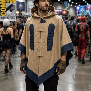 May include: A tan hooded poncho with navy blue trim and accents. The poncho has a button closure and is worn with brown gloves and black trousers. The background shows a convention hall with people and signage.