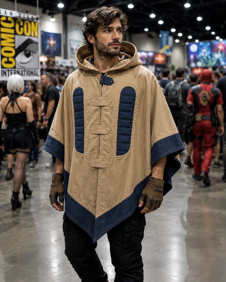 May include: A tan hooded poncho with navy blue trim and accents. The poncho has a button closure and is worn with brown gloves and black trousers. The background shows a convention hall with people and signage.