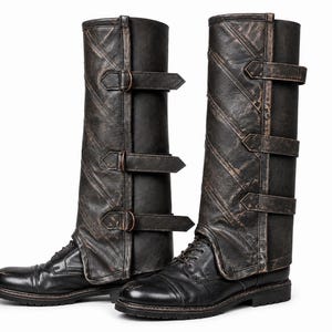 May include: Two black leather spats with a distressed finish, secured with multiple buckles. These spats are designed to be worn over boots, giving a vintage or steampunk look. The boots are black, with a classic lace-up design.