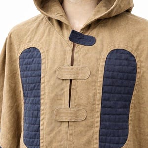 May include: A tan hooded poncho with navy blue quilted panels and accents. The poncho has a button closure and a hood. The design includes navy blue fabric on the cuffs and a navy blue accent on the hood.