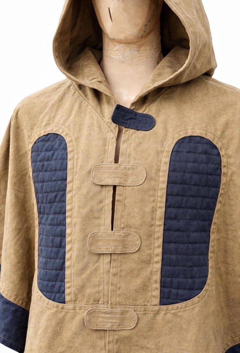 May include: A tan hooded poncho with navy blue quilted panels and accents. The poncho has a button closure and a hood. The design includes navy blue fabric on the cuffs and a navy blue accent on the hood.