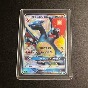 Könnte beinhalten: Eine Charizard GX Sammelkarte in einer Schutzhülle. Die Karte zeigt eine blaue und orangefarbene Charizard-Illustration mit japanischem Text und numerischen Werten. Die Karte ist vor einem dunklen Hintergrund.