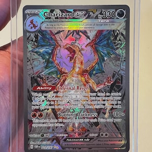 May include: A Charizard ex Tera trading card with 330 HP, featuring a crystalline artwork of the dragon-like creature. The card displays the Infernal Reign ability and Burning Darkness attack, with a dark, shattered-glass background effect.
