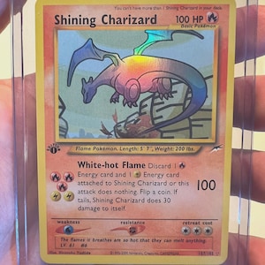 May include: A Shining Charizard trading card featuring a holographic dragon illustration, 100 HP, and the White-hot Flame attack. The card includes text details like Flame Pokémon, 5' 7" length, 200 lbs weight, and the 1st Edition stamp.
