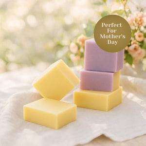 Op de afbeelding: Gele en lavendelkleurige vierkante zepen zijn gestapeld en gerangschikt op een witte doek. De zepen zijn in een stapel van drie en twee individuele zepen. De tekst "Perfect For Mother's Day" staat in een gouden cirkel.