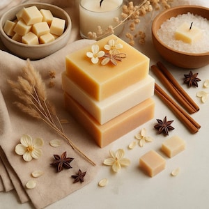 May include: A stack of three square soaps in shades of beige and tan, decorated with small white flowers and a bundle of dried stems. Additional soaps, star anise, and cinnamon sticks are arranged around the stack. A bowl of cubes and a bowl of bath salts are also present.