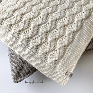 May include: Cream-colored knitted blanket with a textured, repeating leaf pattern. The blanket is folded, revealing a corner with a small tag that reads "happyknitball". The blanket is resting on a gray patterned surface.