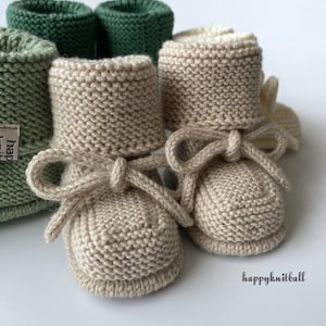 May include: Close-up of knitted baby booties in beige with matching bows. Several other knitted items in shades of green are visible in the background. The booties are soft and appear handmade. The overall theme is baby clothing.