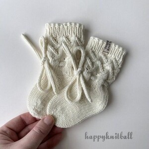 May include: A pair of cream-colored knitted socks featuring a textured cable knit pattern on the cuffs and decorative tied bows on the sides. A small fabric tag with the text "happy knitball" is attached to the cuff of one sock.