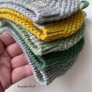 May include: A stack of five folded knit hats in shades of light gray, mustard yellow, sage green, dark forest green, and medium gray. Each hat features a small rectangular fabric tag with the text happyknitball printed in black ink.