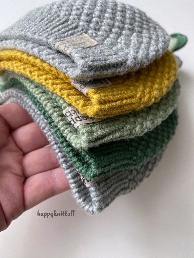 May include: A stack of five folded knit hats in shades of light gray, mustard yellow, sage green, dark forest green, and medium gray. Each hat features a small rectangular fabric tag with the text happyknitball printed in black ink.
