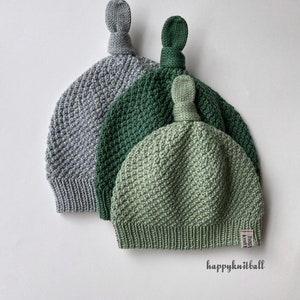 May include: Three knit hats in gray, dark green, and light green, each featuring a textured stitch pattern, a ribbed brim, and a top knot. A small white tag with the text "happyknitball" is attached to the light green hat, and the text "happyknitball" is printed below.