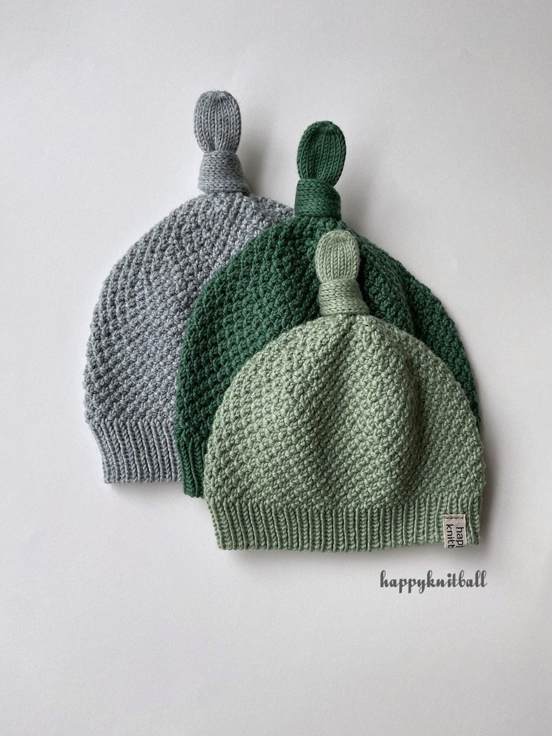 May include: Three knit hats in gray, dark green, and light green, each featuring a textured stitch pattern, a ribbed brim, and a top knot. A small white tag with the text "happyknitball" is attached to the light green hat, and the text "happyknitball" is printed below.