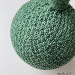May include: A spherical object made of sage green yarn with a dense, textured crochet stitch pattern. A small knotted loop extends from the top of the sphere. The text "happyknitball" is printed in a small, dark font in the lower right corner.
