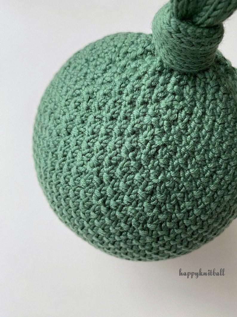 May include: A spherical object made of sage green yarn with a dense, textured crochet stitch pattern. A small knotted loop extends from the top of the sphere. The text "happyknitball" is printed in a small, dark font in the lower right corner.