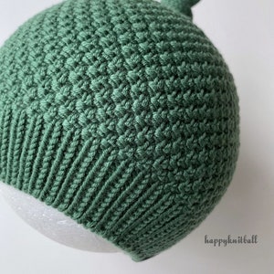 May include: A sage green knit hat featuring a textured stitch pattern on the main body, a ribbed brim, and a small decorative knot at the top. The text "happyknitball" is printed in the lower right corner of the image.