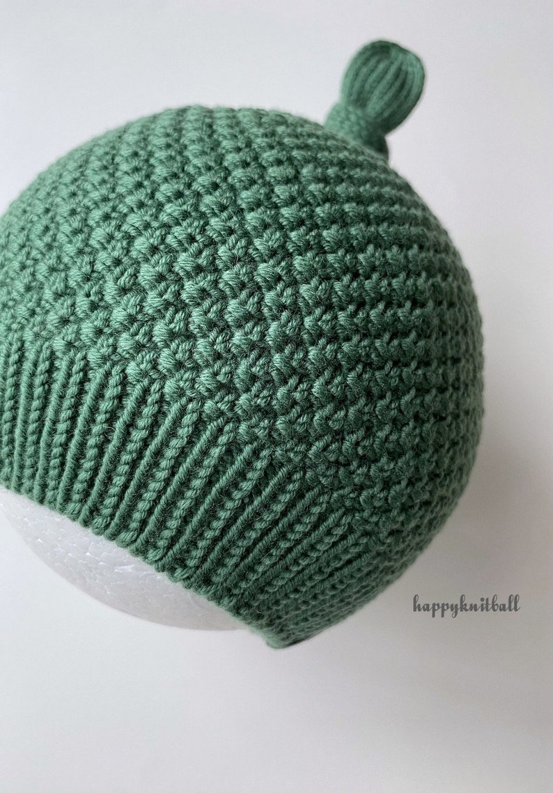 May include: A sage green knit hat featuring a textured stitch pattern on the main body, a ribbed brim, and a small decorative knot at the top. The text "happyknitball" is printed in the lower right corner of the image.