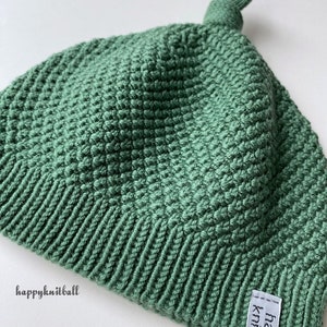 May include: A sage green knit hat featuring a textured stitch pattern, a ribbed brim, and a small knot at the top. A white fabric tag with the black text happyknitball is sewn onto the brim, and the text happyknitball is printed on the side.