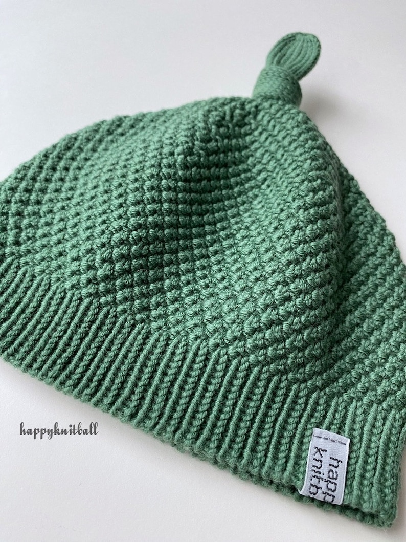 May include: A sage green knit hat featuring a textured stitch pattern, a ribbed brim, and a small knot at the top. A white fabric tag with the black text happyknitball is sewn onto the brim, and the text happyknitball is printed on the side.