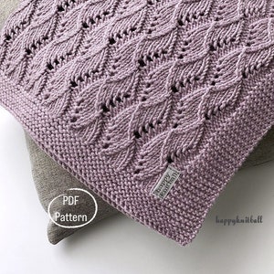 Op de afbeelding: Een close-up van een zacht, paars gebreid deken met een delicaat, kantpatroon. Het deken is gevouwen en ligt op een lichtbruin oppervlak. De tekst "PDF Pattern" is zichtbaar op het oppervlak. De tekst "happy knitball" is zichtbaar op een wit label dat aan het deken is bevestigd.