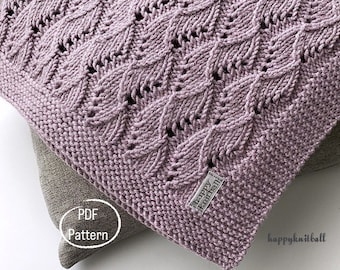 Könnte beinhalten: Nahaufnahme einer weichen, lila gestrickten Decke mit einem zarten, durchbrochenen Muster. Die Decke ist gefaltet und liegt auf einer hellbraunen Oberfläche. Der Text "PDF Pattern" ist auf der Oberfläche sichtbar. Der Text "happy knitball" ist auf einem weißen Etikett sichtbar, das an der Decke befestigt ist.