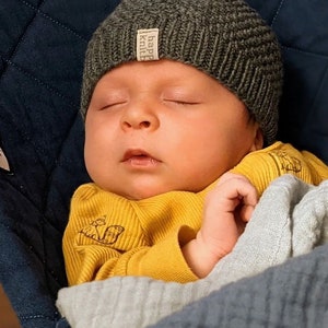 May include: A dark gray ribbed knit beanie with a small beige fabric tag featuring the text "happy knitb" in black. The beanie is worn by a sleeping infant dressed in a mustard yellow long-sleeved ribbed shirt with a small black line drawing.